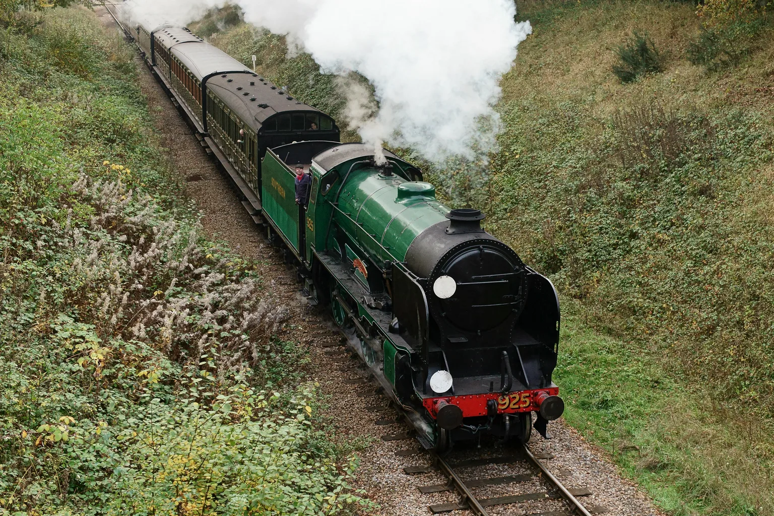 A green steam locomotive numbered 925 climbing through a cutting on the Bluebell Railway with steam rising.