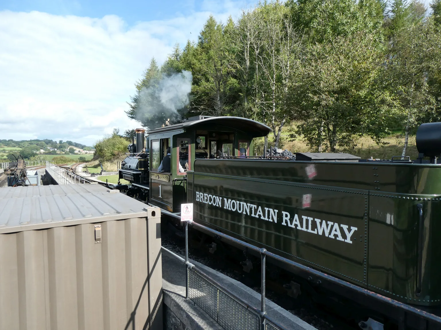A green steam locomotive and tender lettered Brecon Mountain Railway at Pant station, trees and hills behind