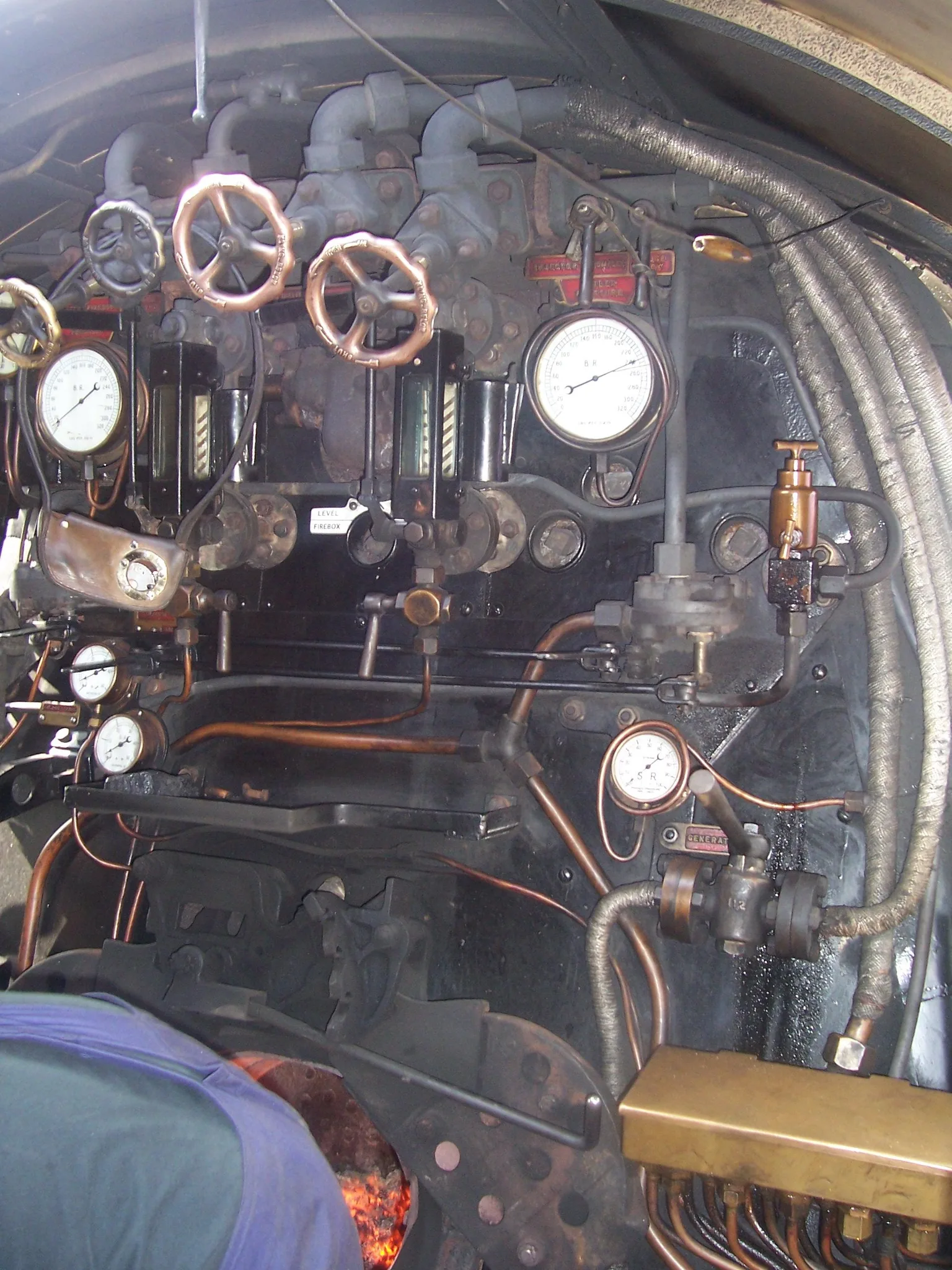 The inside of a steam locomotive cab: brass and copper valves, round pressure gauges and pipes above the glow of the firebox