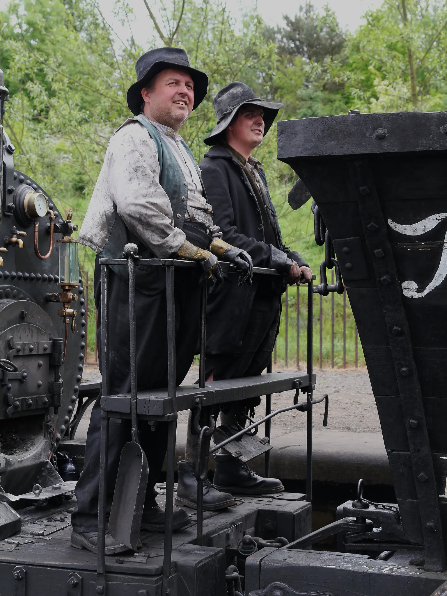 Two crew in work clothes and hats standing on the footplate of an early steam locomotive in green countryside