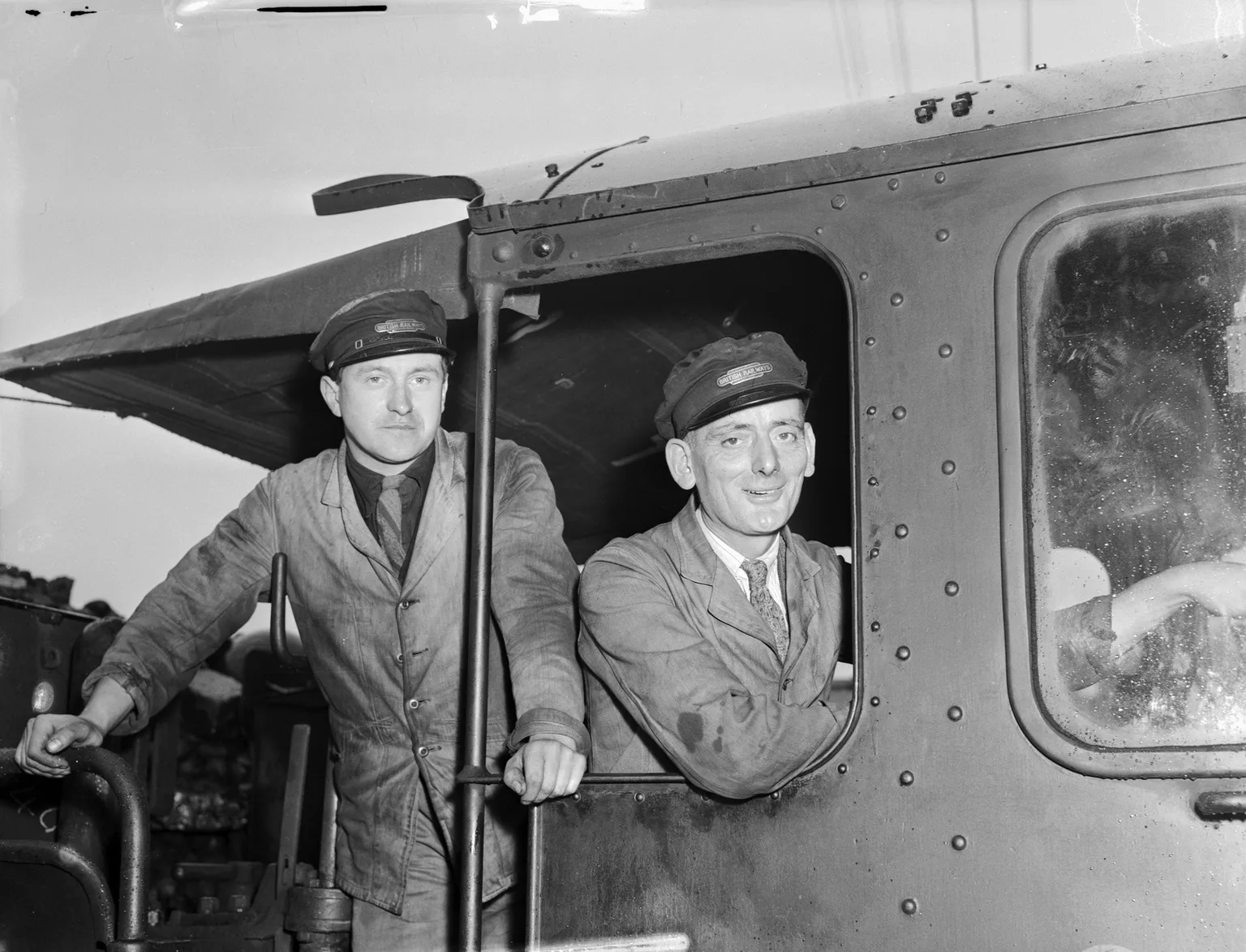 A black and white photograph of two railwaymen in caps leaning out of the cab of a steam locomotive, smiling.