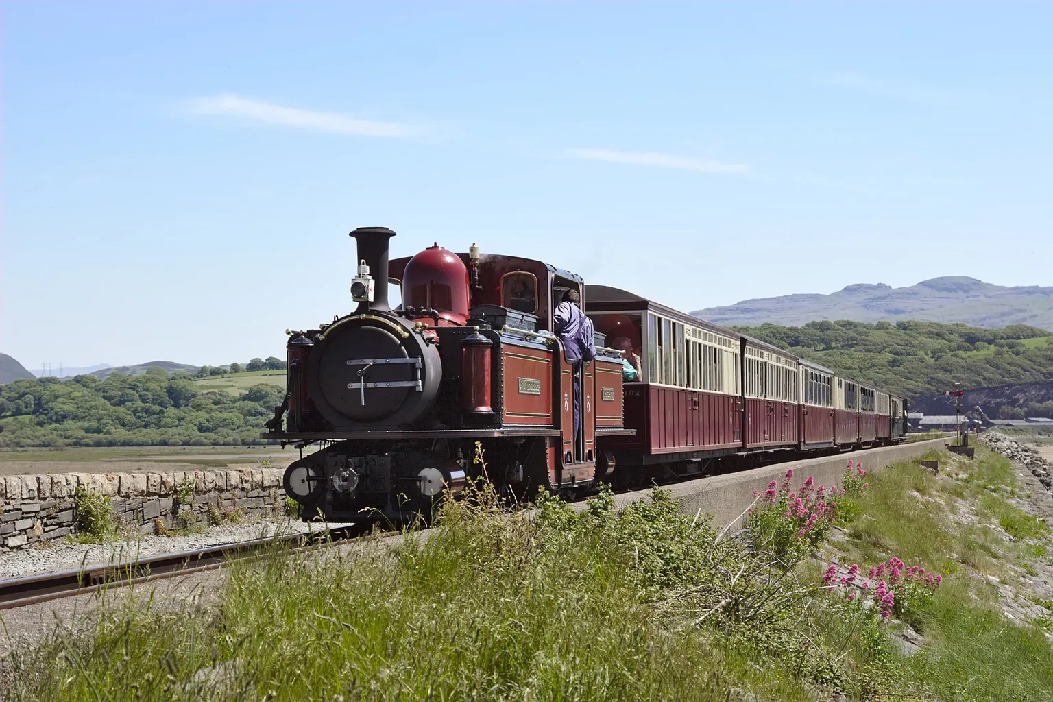 A red narrow gauge steam locomotive and carriages crossing the Cob at Porthmadog, hills beyond.