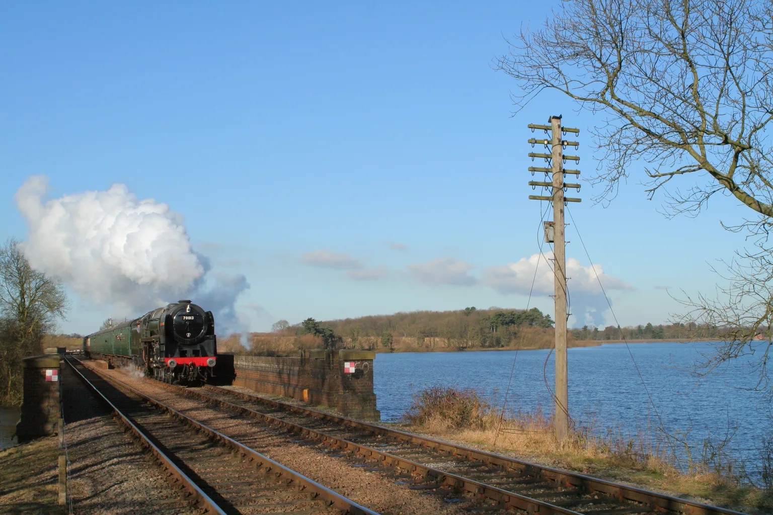 A steam express crossing the embankment by Swithland reservoir under a blue sky, a telegraph pole in the foreground.