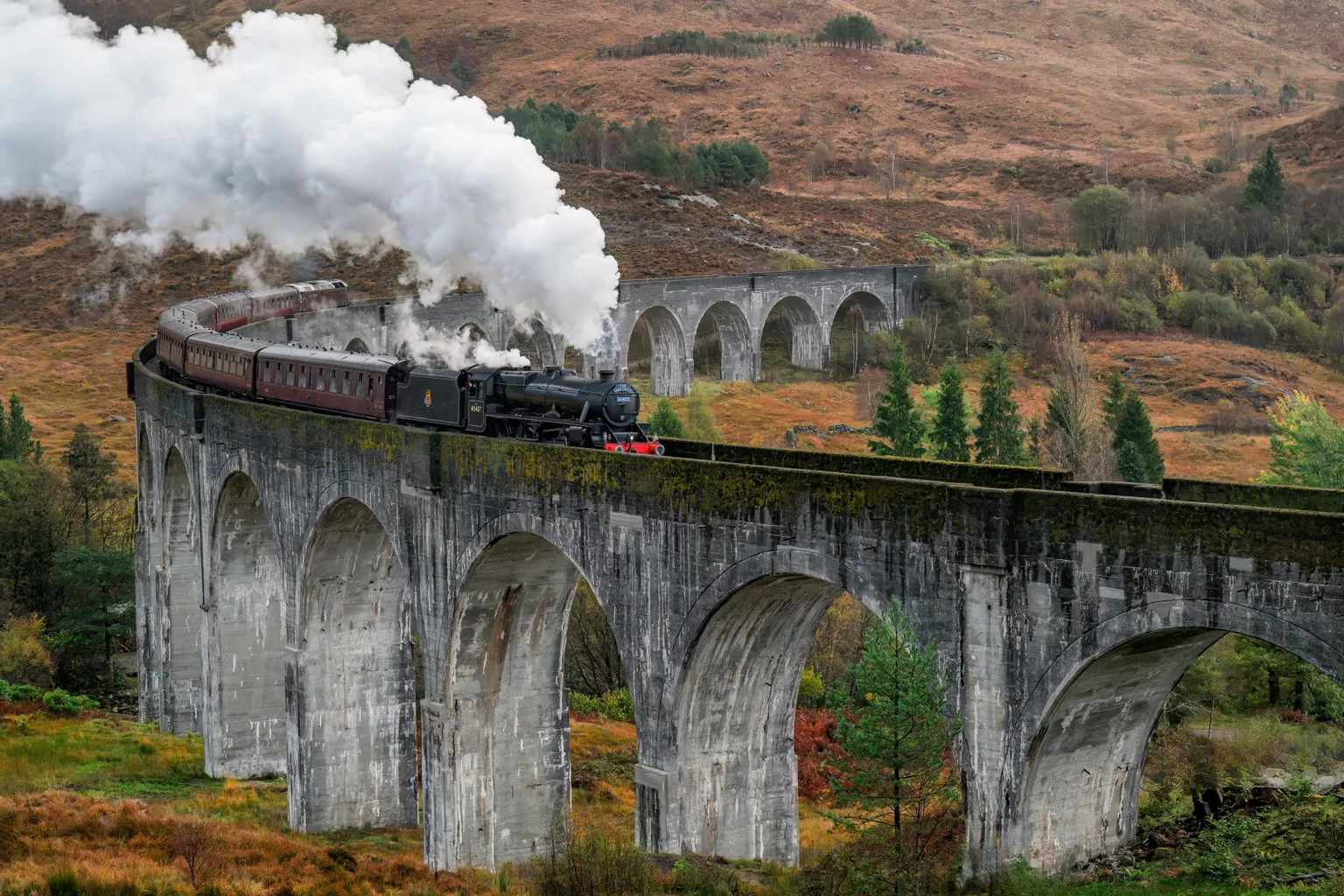 A black steam locomotive numbered 45407 hauling maroon carriages across the concrete arches of the Glenfinnan Viaduct, a big white plume of steam behind it