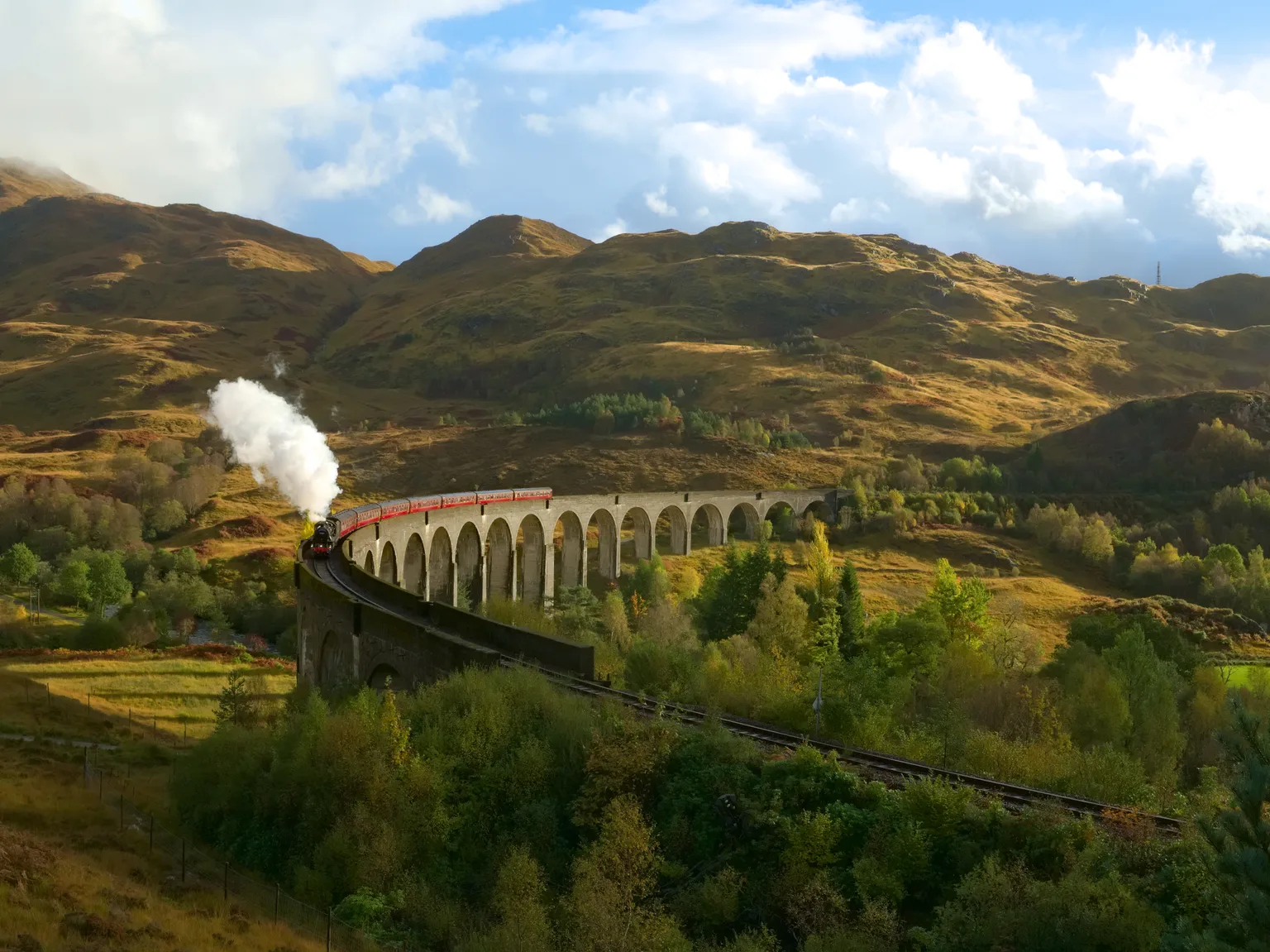 A steam train with red carriages curving across the Glenfinnan Viaduct, autumn hills and a bright cloudy sky behind.