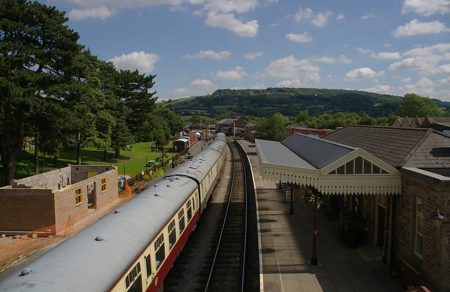 A long rake of carriages at Winchcombe station seen from the footbridge, wooded hills beyond
