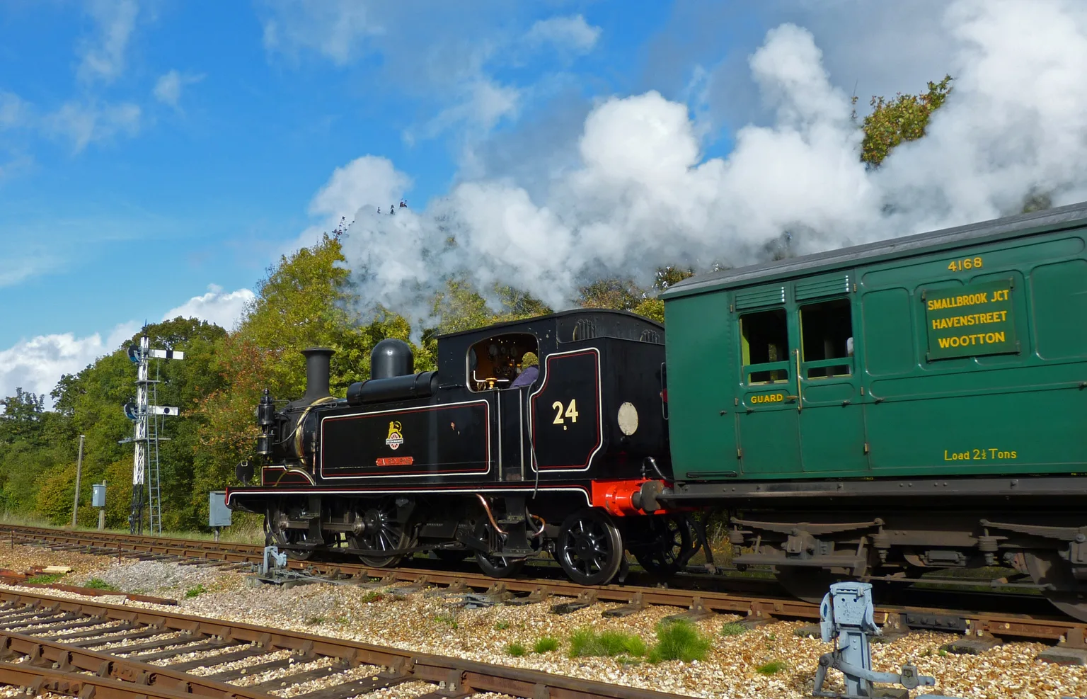 A black tank engine numbered 24 beside a green carriage under a blue sky at Havenstreet, steam drifting.