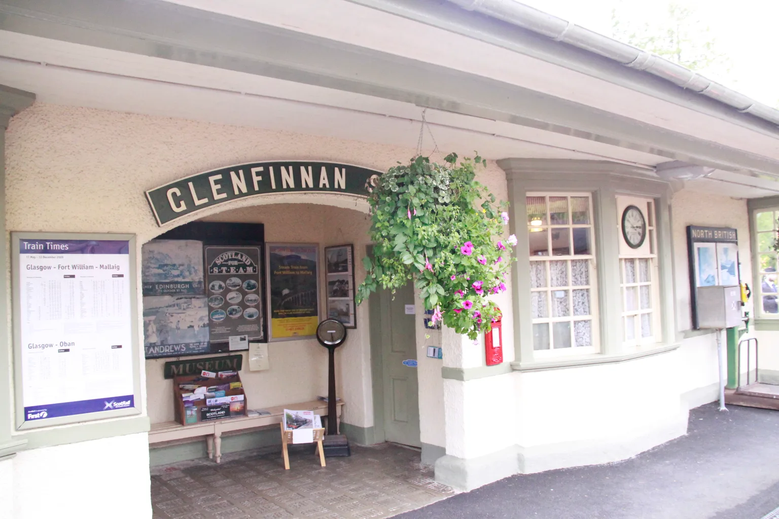 The cream station building at Glenfinnan with its arched sign, timetable boards and hanging baskets
