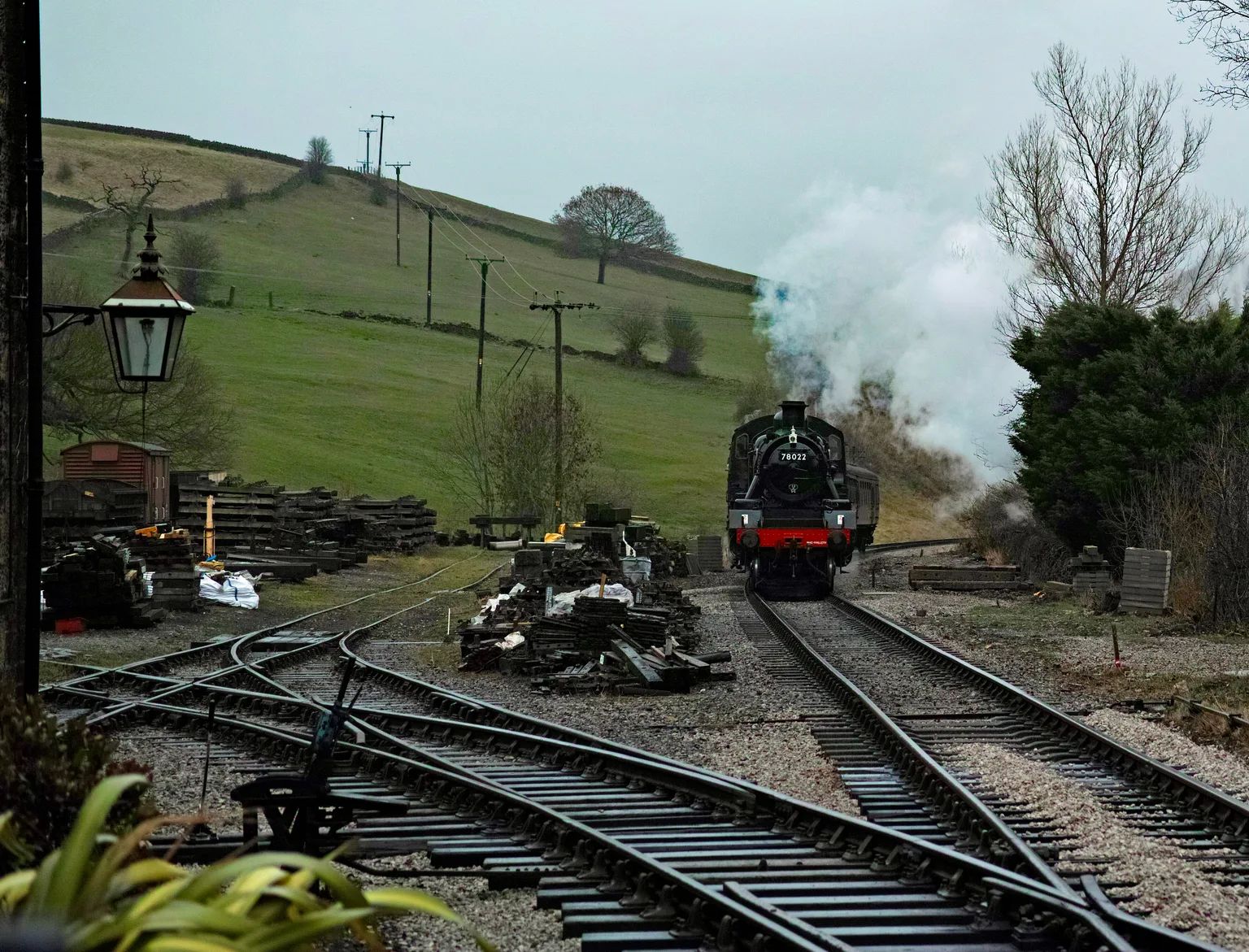 A steam locomotive running into Oakworth station past the yard, green hills behind.