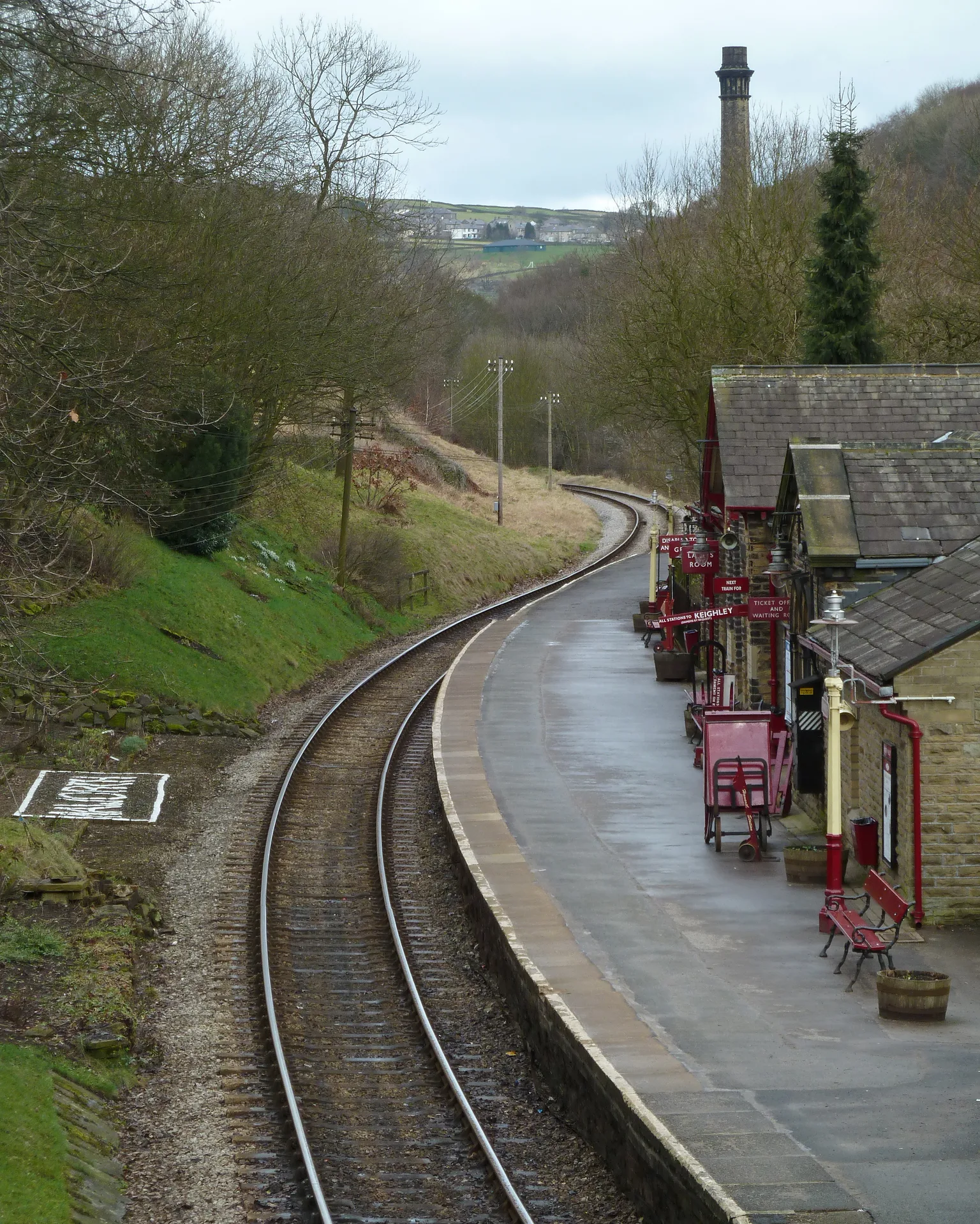 A curving platform at Haworth with a stone station building, red lamps and a chimney on the hill