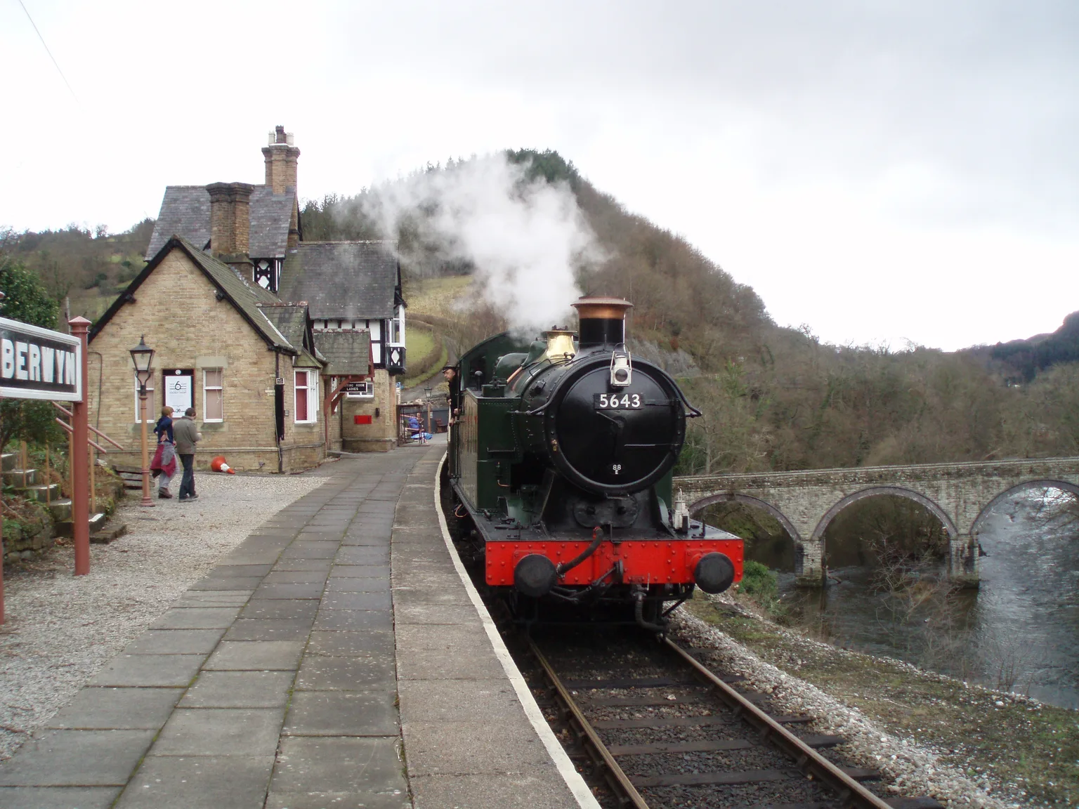 A green steam tank engine arriving at Berwyn station beside a stone building, a viaduct over the River Dee behind.