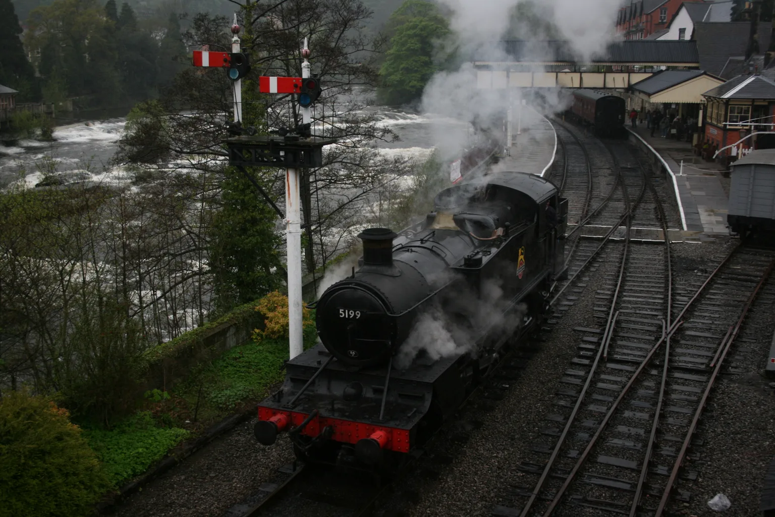 A black steam locomotive beside signals at Llangollen station, the River Dee rushing past below