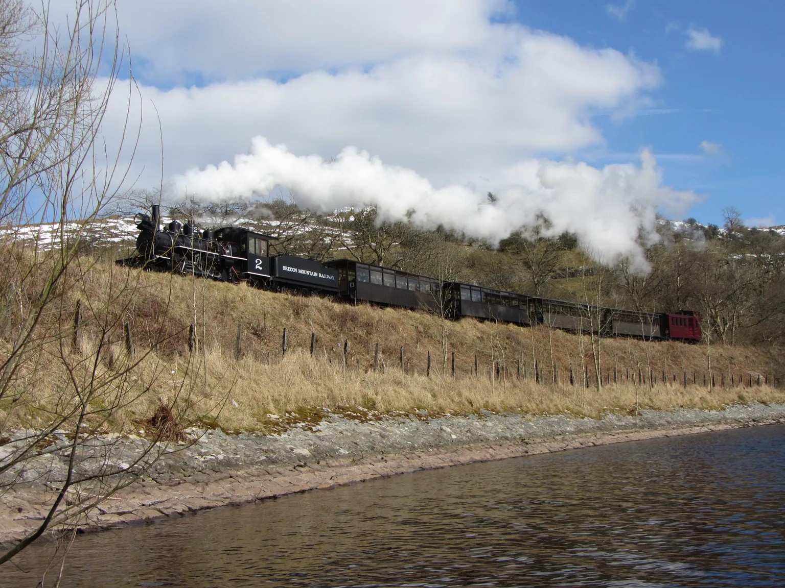 A small black steam locomotive and carriages running beside a reservoir below snowy hills on the Brecon Mountain Railway