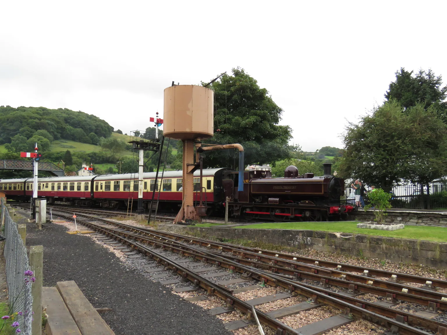 Maroon and cream carriages and a small steam engine beside a water tower in the yard at Buckfastleigh