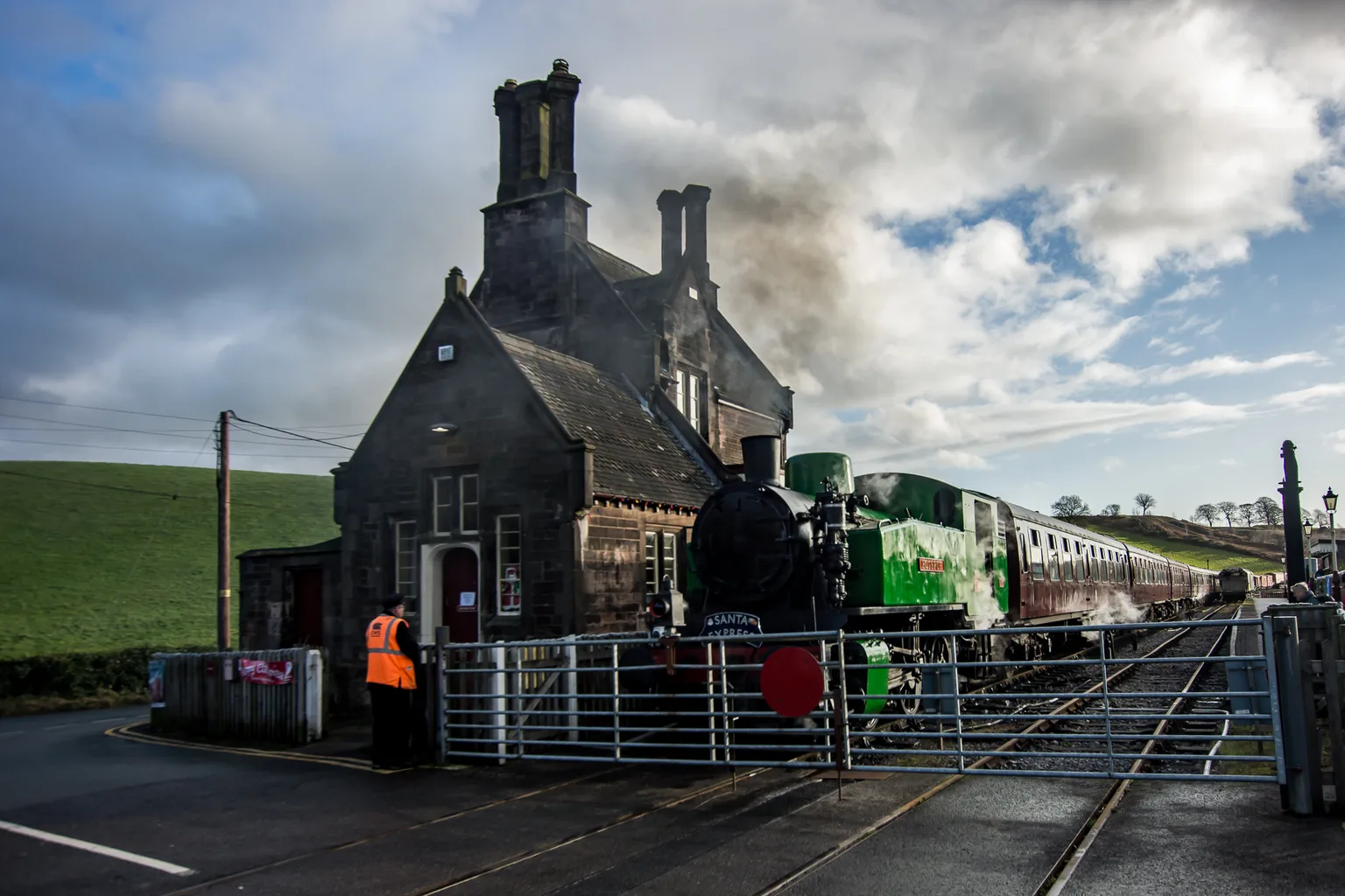 A green steam locomotive and carriages beside the stone station building at Cheddleton, behind a level crossing gate
