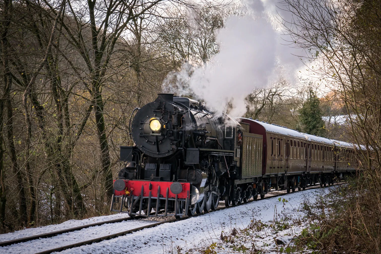 A black steam locomotive with its headlamp lit hauling maroon carriages through a snowy wood on the Churnet Valley Railway.