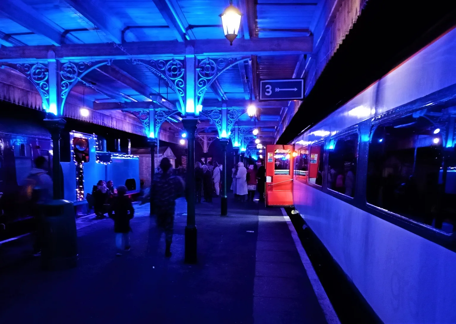 A station platform at night lit in blue, families in coats walking beside a carriage with a red door open and a small engine strung with lights on the left