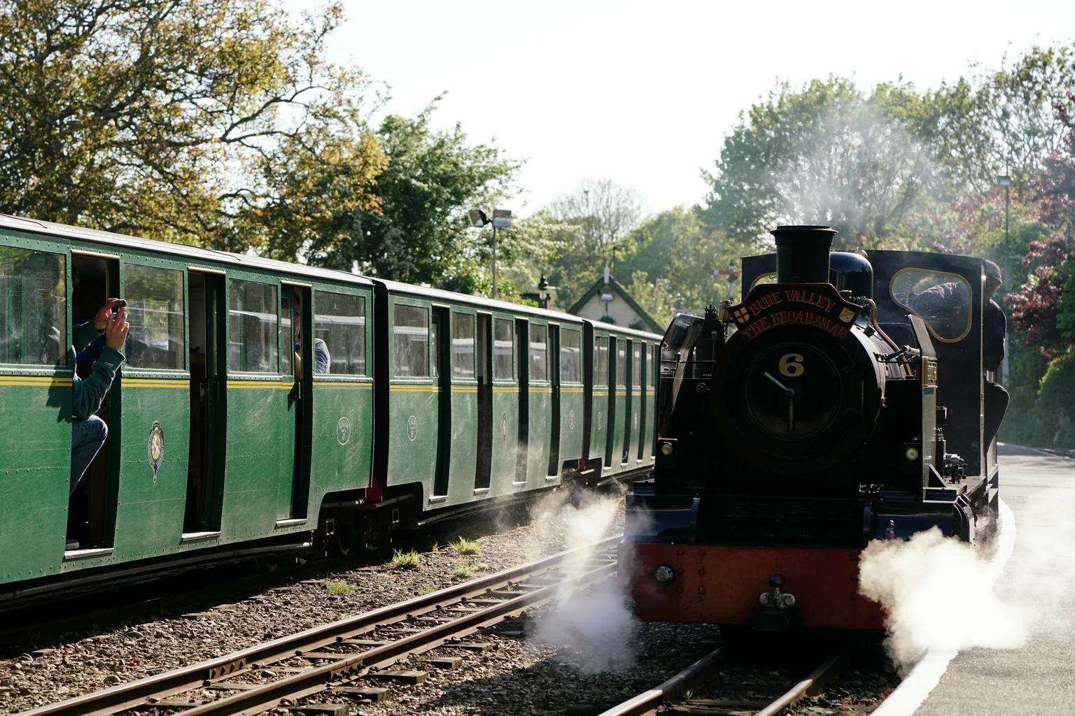 A small black steam locomotive numbered 6 in steam beside a long train of green miniature carriages at a station, trees behind.