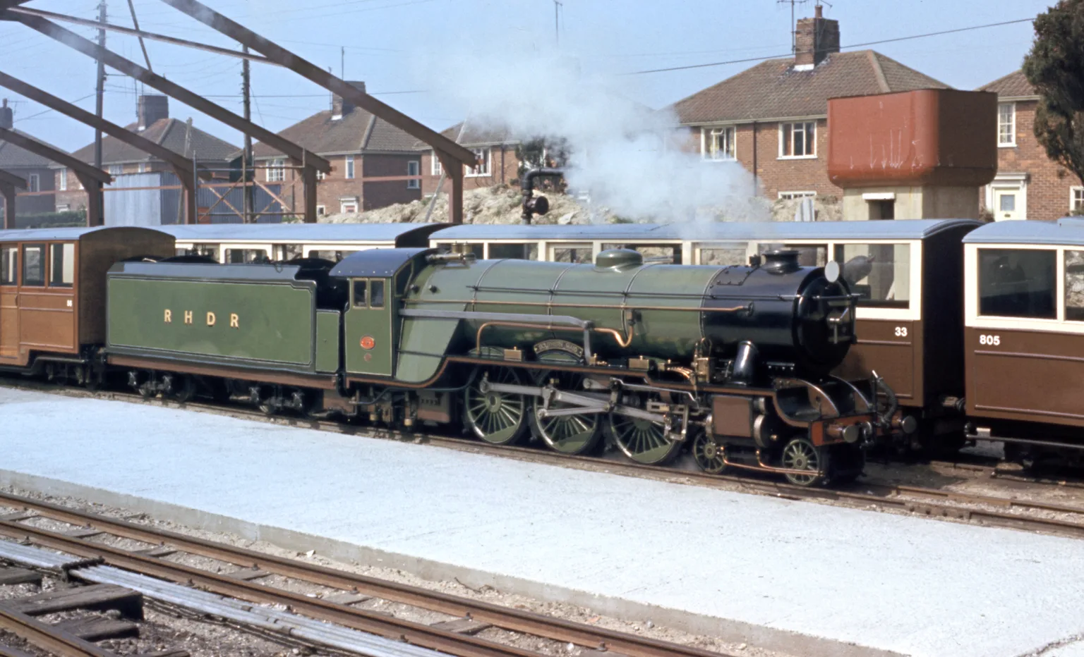 A green miniature steam locomotive lettered RHDR with brown carriages at New Romney station, houses behind