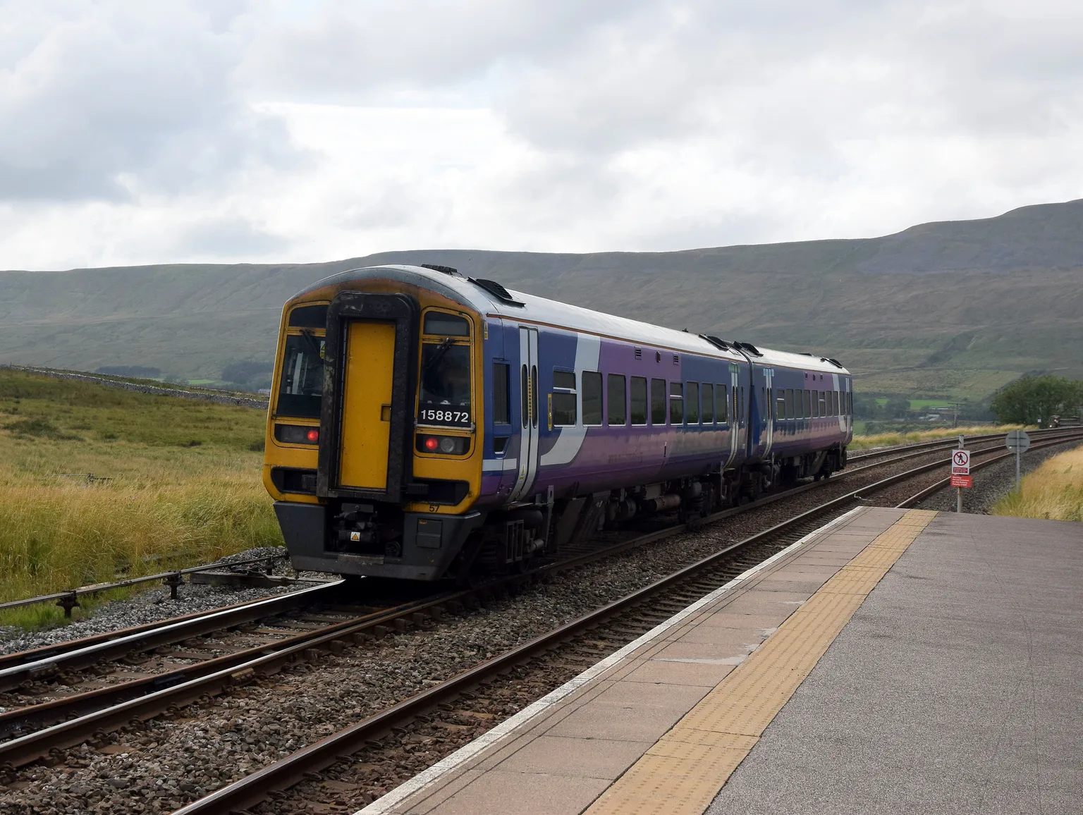 A blue and purple Northern train standing at the platform of Ribblehead station, with green fells behind under a grey sky