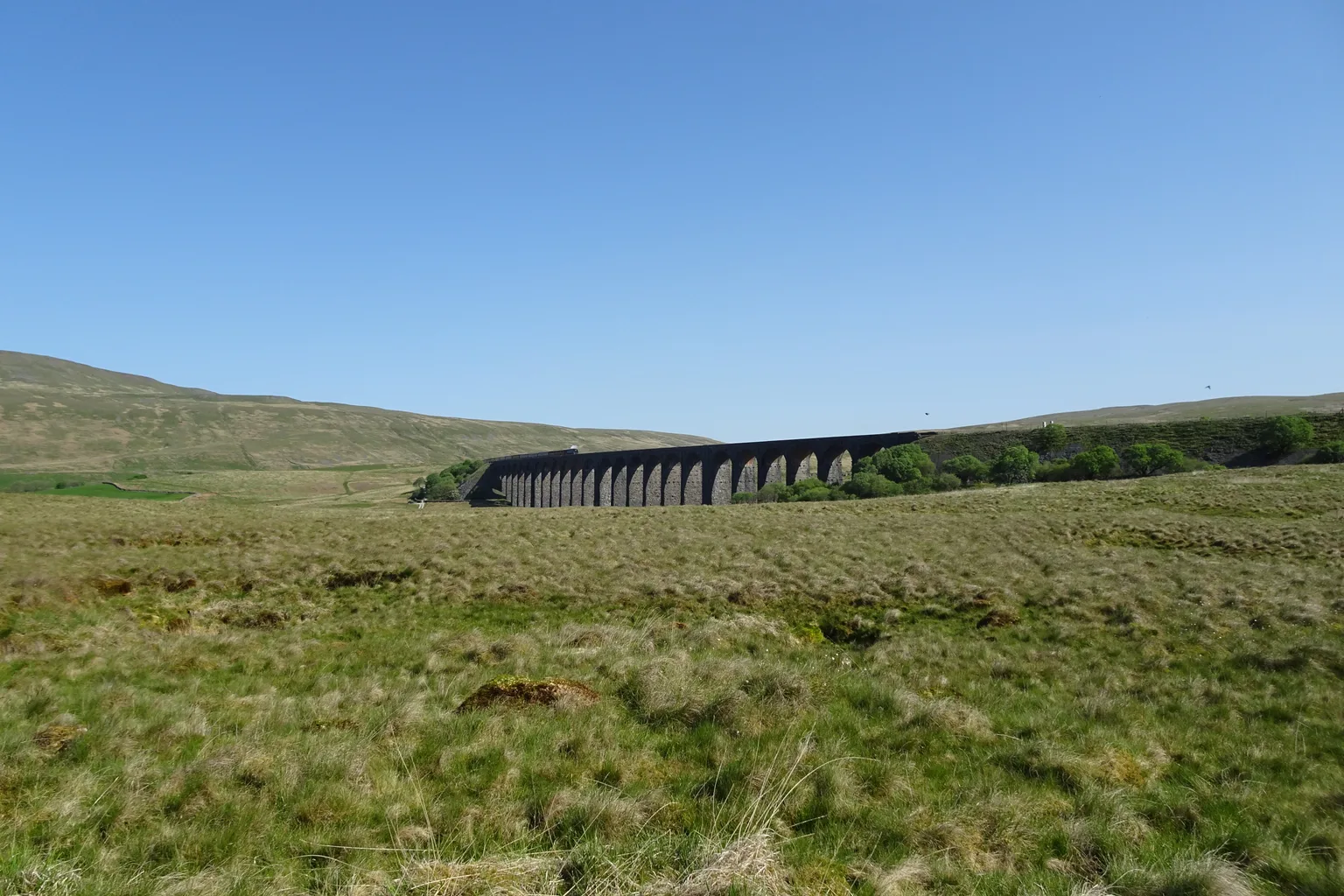 The long row of stone arches of the Ribblehead Viaduct across open moorland under a clear blue sky, a train on top.