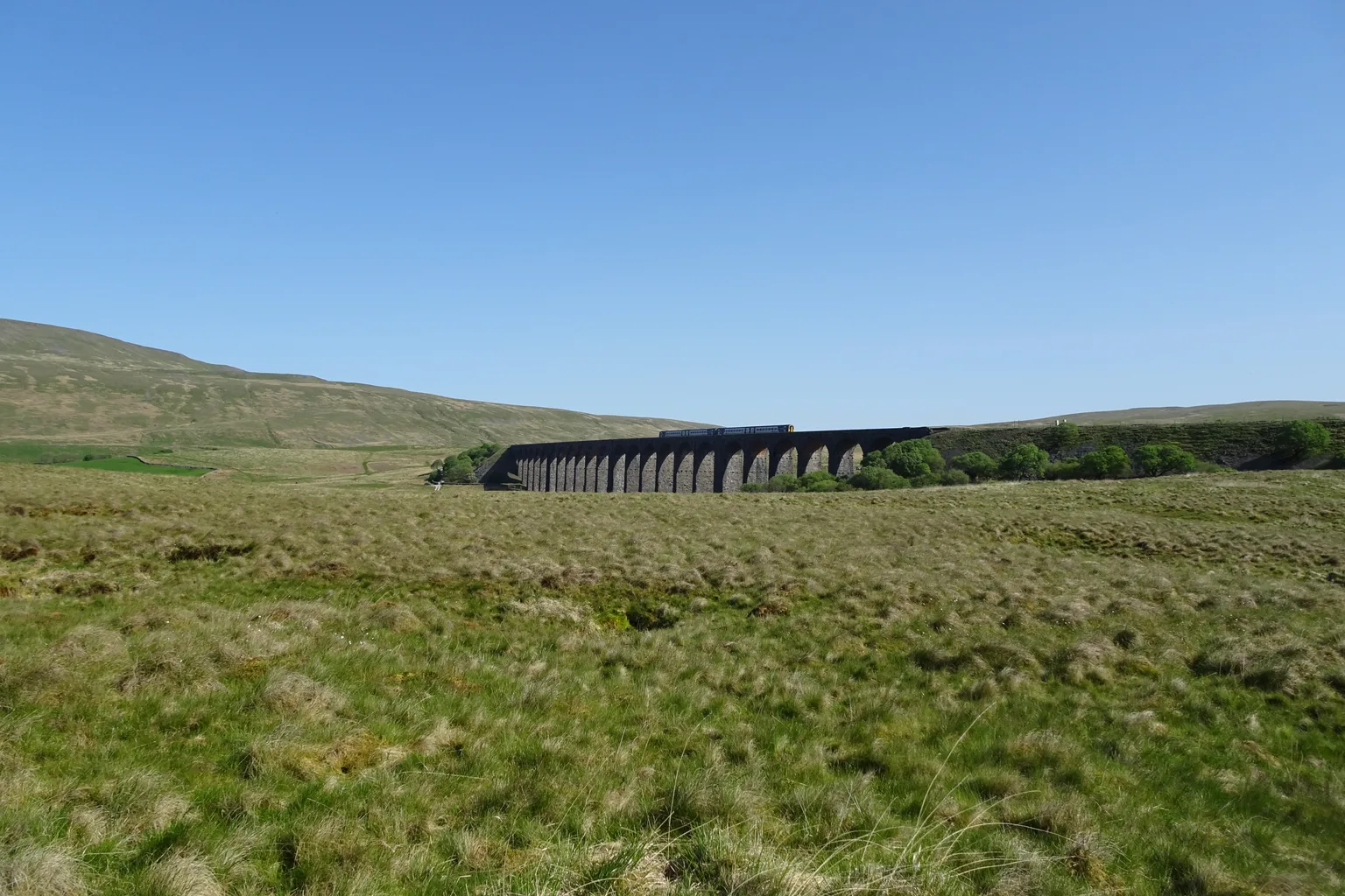 The Ribblehead Viaduct seen far across a rough grassy moor, its arches dark against a clear blue sky