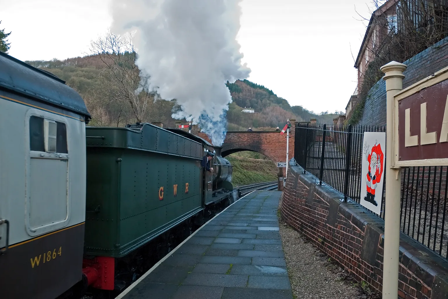 A green tank engine with GWR on its side pulling away from Llangollen station, steam rising, a Santa poster on the platform fence