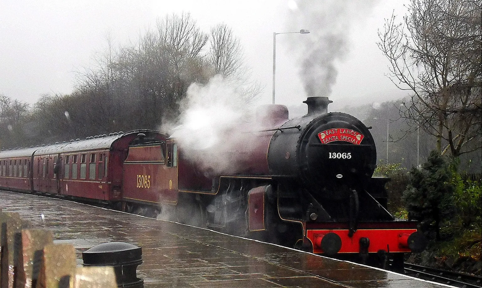 A black and maroon steam locomotive numbered 13065 with a headboard, leading maroon carriages through rain at Rawtenstall station.