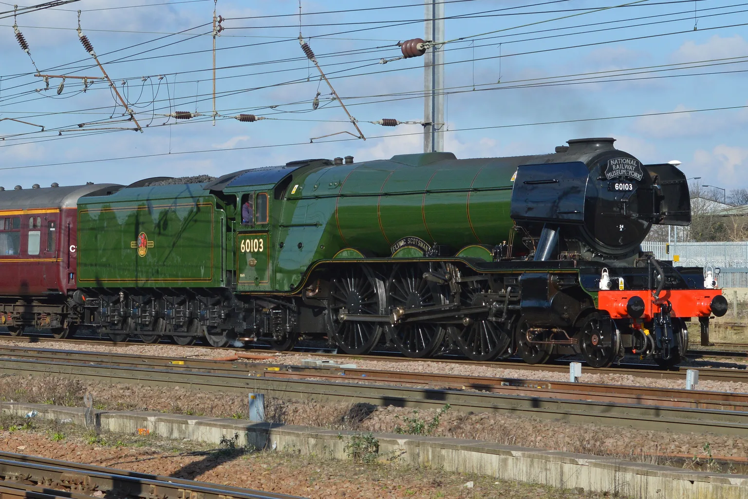 Flying Scotsman, a green steam locomotive numbered 60103 with a National Railway Museum York headboard, running beneath overhead wires with maroon carriages.