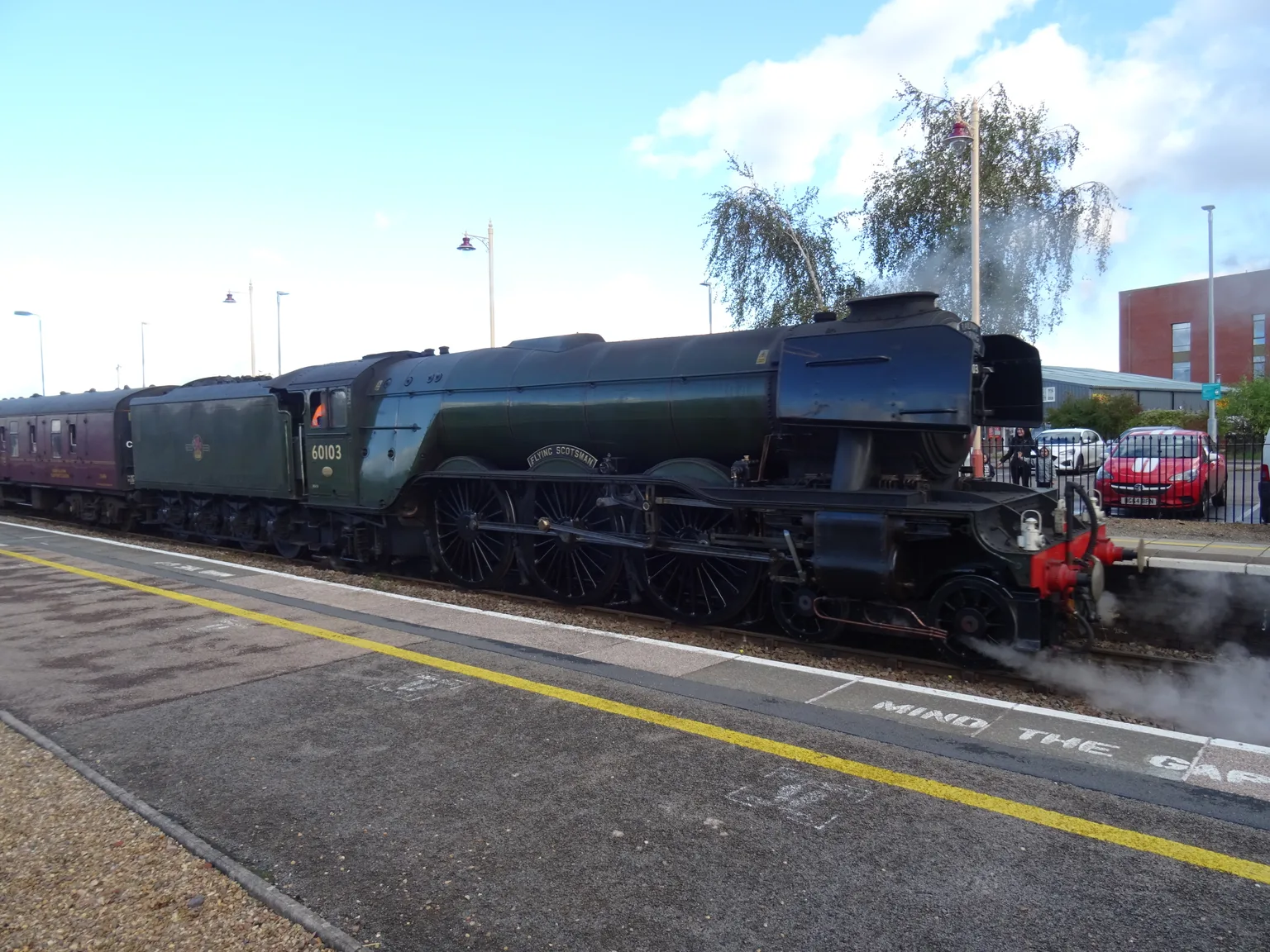 Flying Scotsman in dark green, numbered 60103, standing at a station platform with maroon carriages behind and a red car beyond the railings