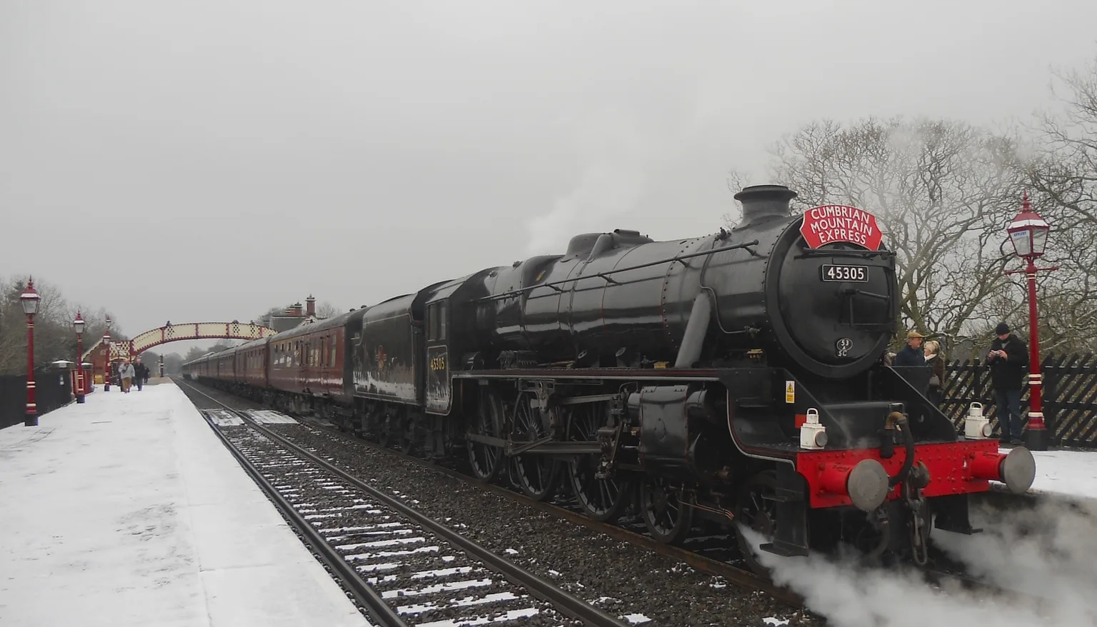Black steam locomotive 45305 with a red Cumbrian Mountain Express headboard at a snowy station platform beside red lamp posts and a footbridge
