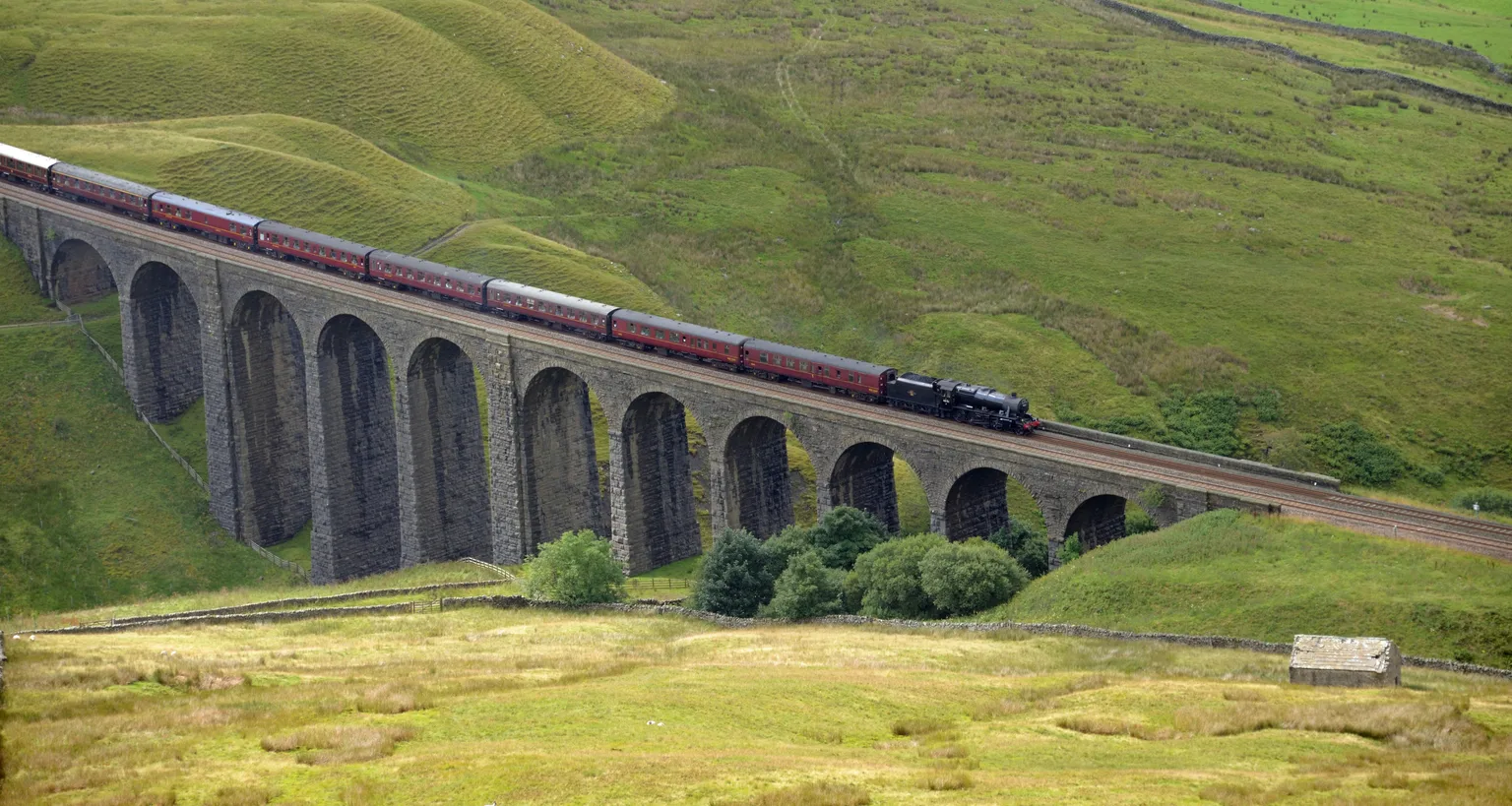 A black steam locomotive hauling maroon carriages across the stone arches of Arten Gill Viaduct below green fells