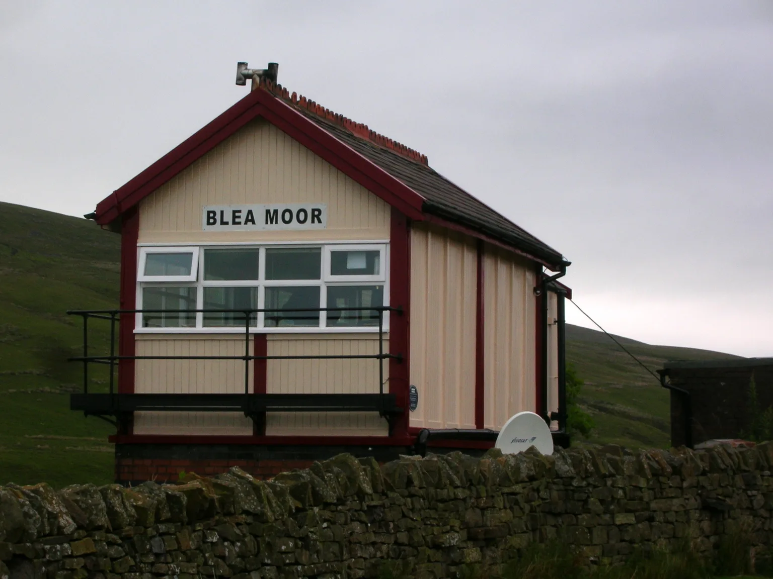 The cream and maroon wooden signal box at Blea Moor behind a dry stone wall, a moor rising behind it
