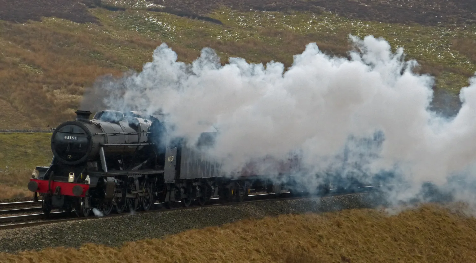 A black steam locomotive working hard through moorland near Garsdale in a great cloud of steam