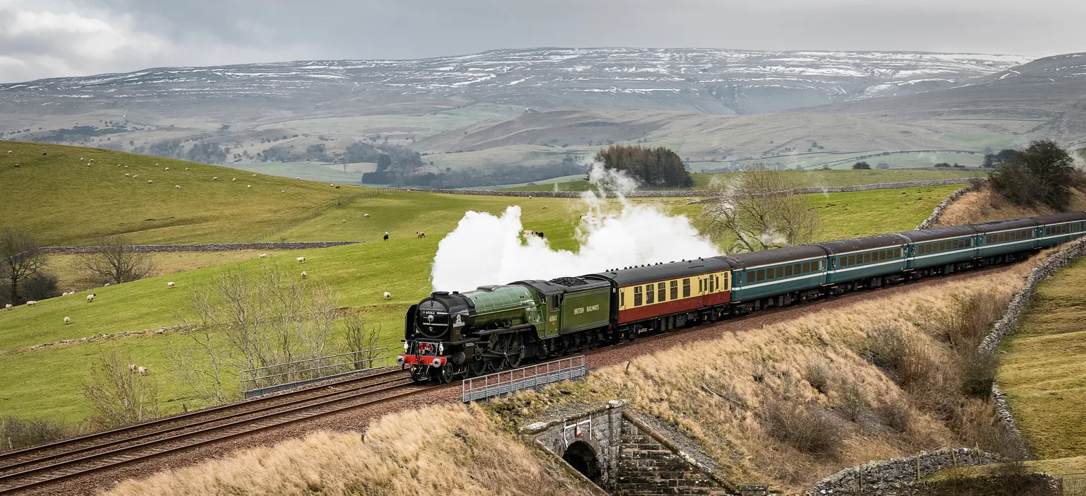 A green express steam locomotive and carriages crossing open fells near Kirkby Stephen, snow on the hills