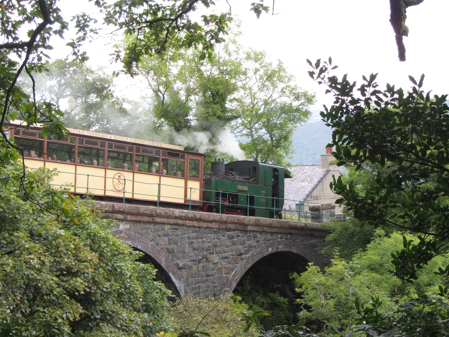 A small steam locomotive and an open-sided carriage on a stone viaduct among trees on the Snowdon Mountain Railway