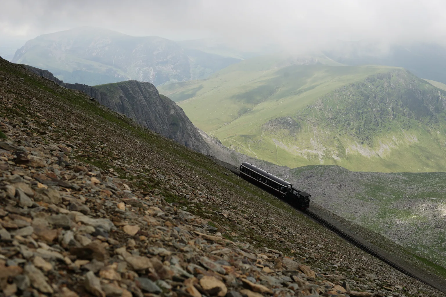 A train climbing a bare stony mountainside on the Snowdon Mountain Railway, a ridge and valley behind