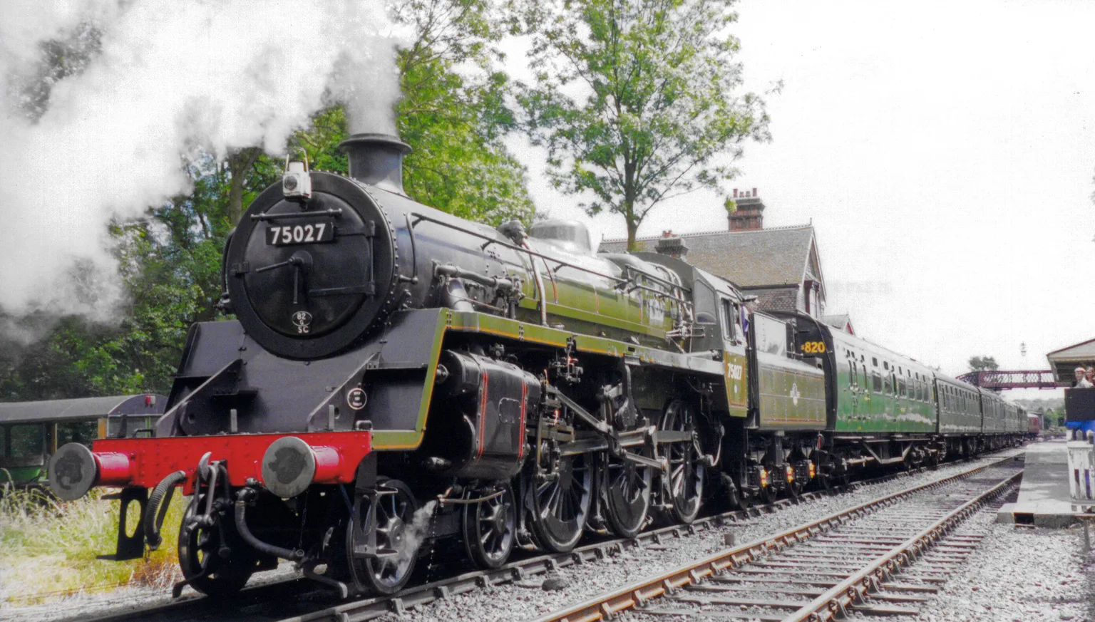 A green British Railways steam locomotive numbered 75027 with its train beside the platform