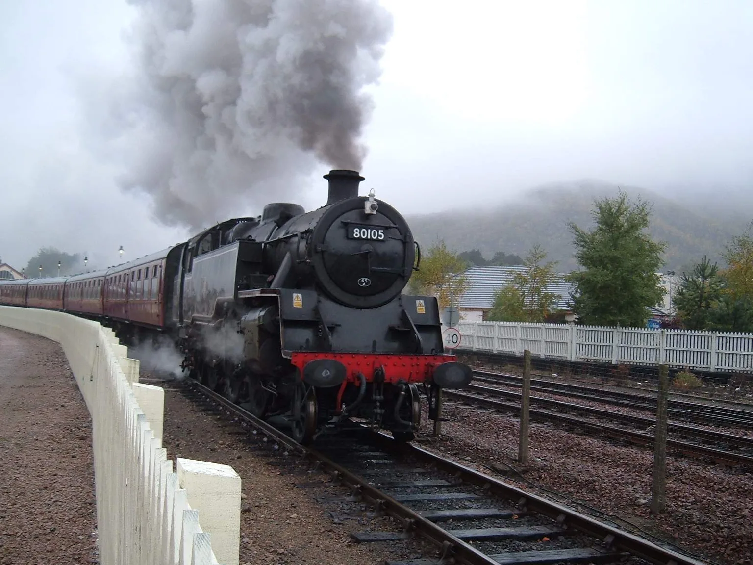 A black steam tank engine pulling away from Aviemore in a plume of smoke, hills under cloud behind.