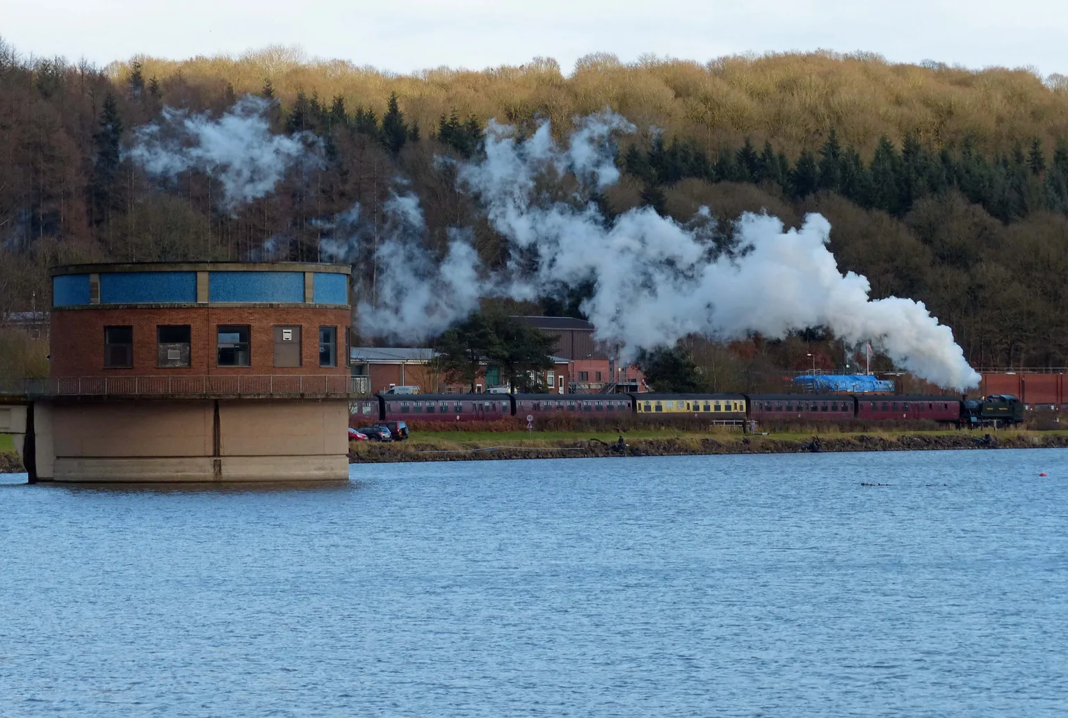 A steam train running beside the reservoir near Trimpley under a wooded bank, the water tower in the foreground.