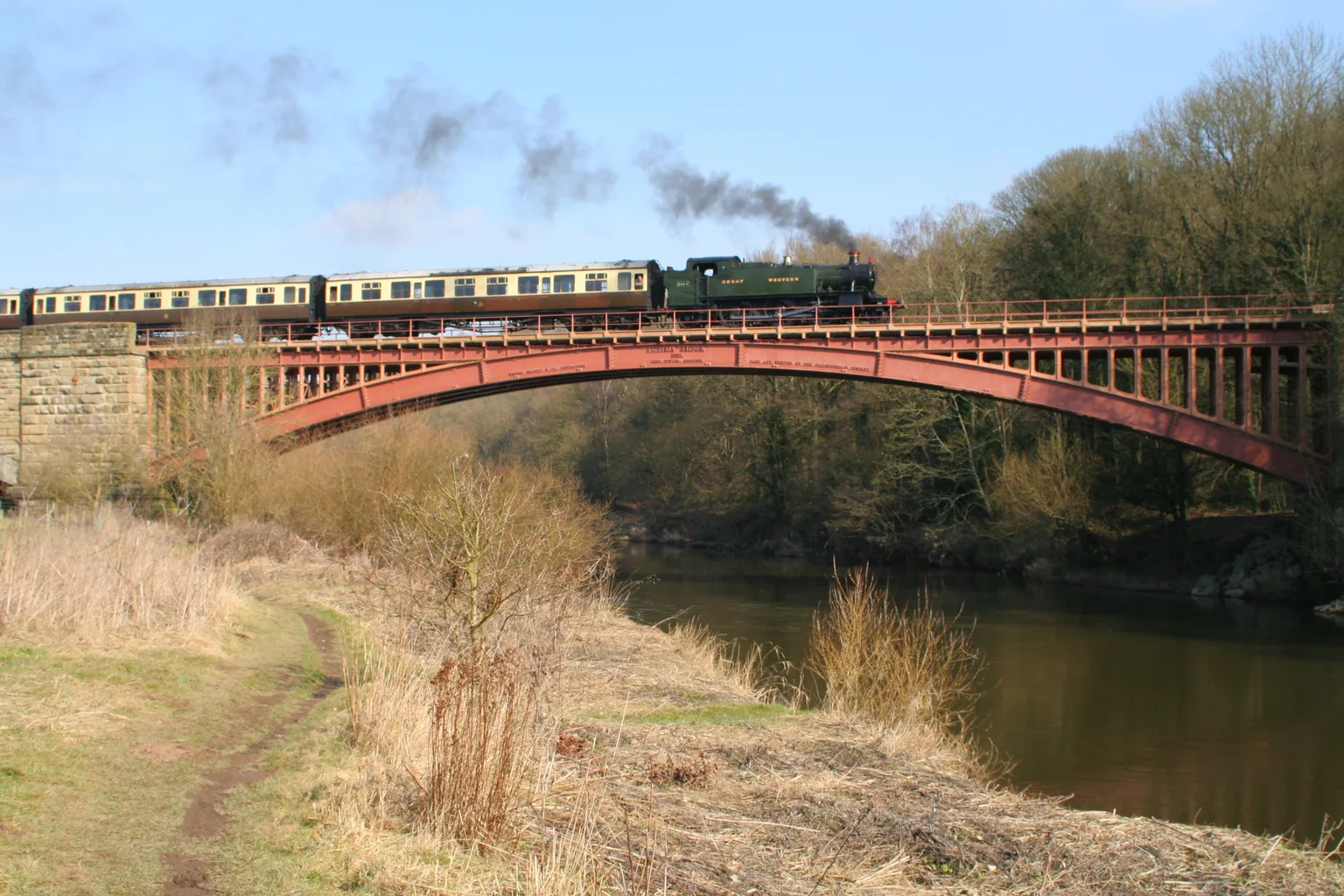 A green tank engine and carriages crossing the red iron arch of the Victoria Bridge over the River Severn