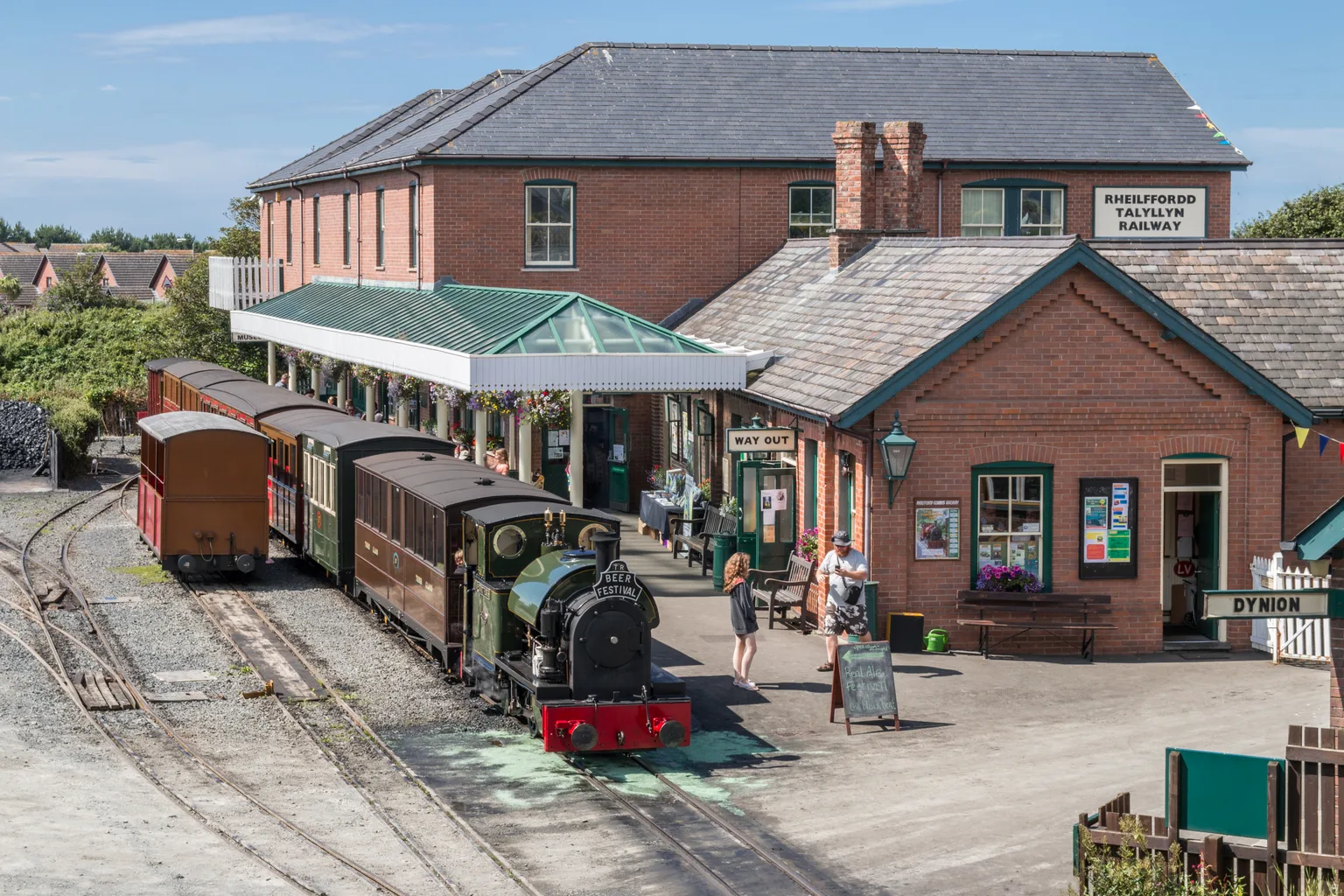 A small green tank engine and brown carriages at Tywyn Wharf station beside the brick station building.