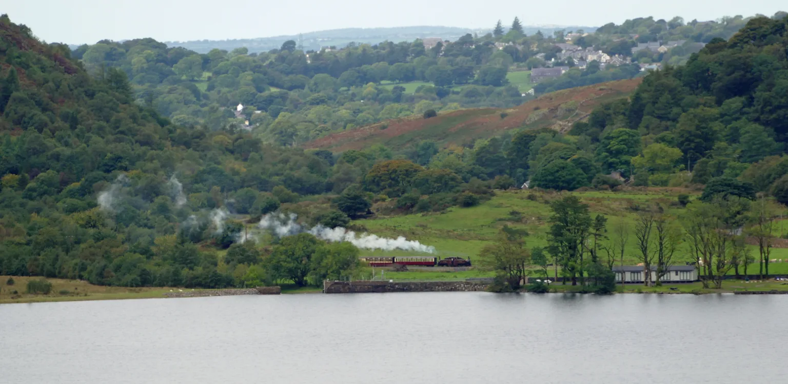 A distant steam train with a white plume running along the far shore of Llyn Cwellyn below wooded hills