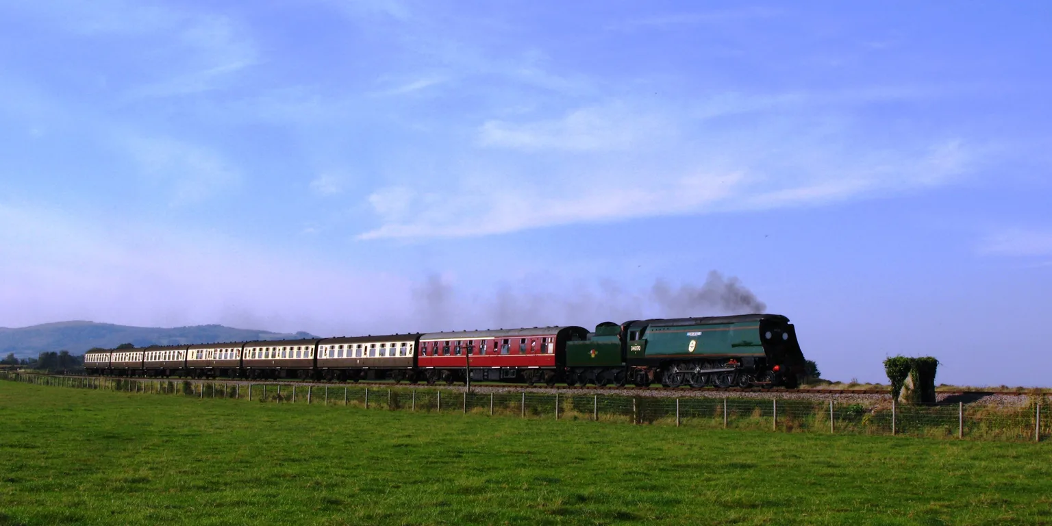 A green Bulleid steam locomotive with a long rake of carriages crossing flat fields near the Somerset coast