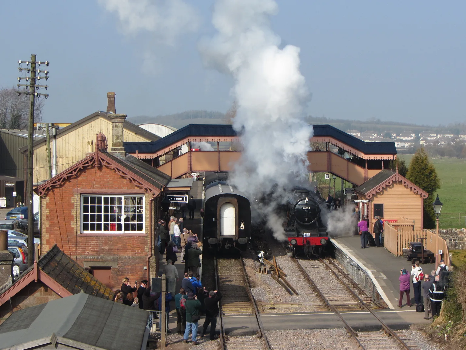 A steam locomotive in a cloud of steam at Williton beside the brick signal box and a footbridge, with a crowd on the platform